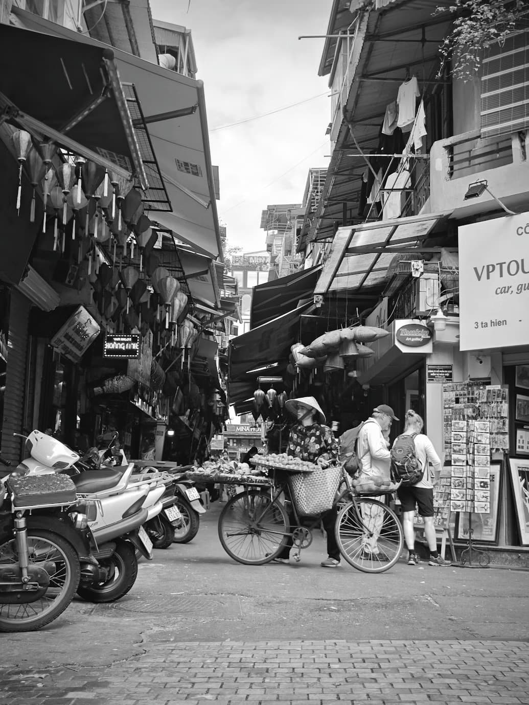 Ruelle du vieux quartier de Hanoï en noir et blanc : une marchande en chapeau conique pousse un vélo chargé de paniers, entre les étals et les motos garées.